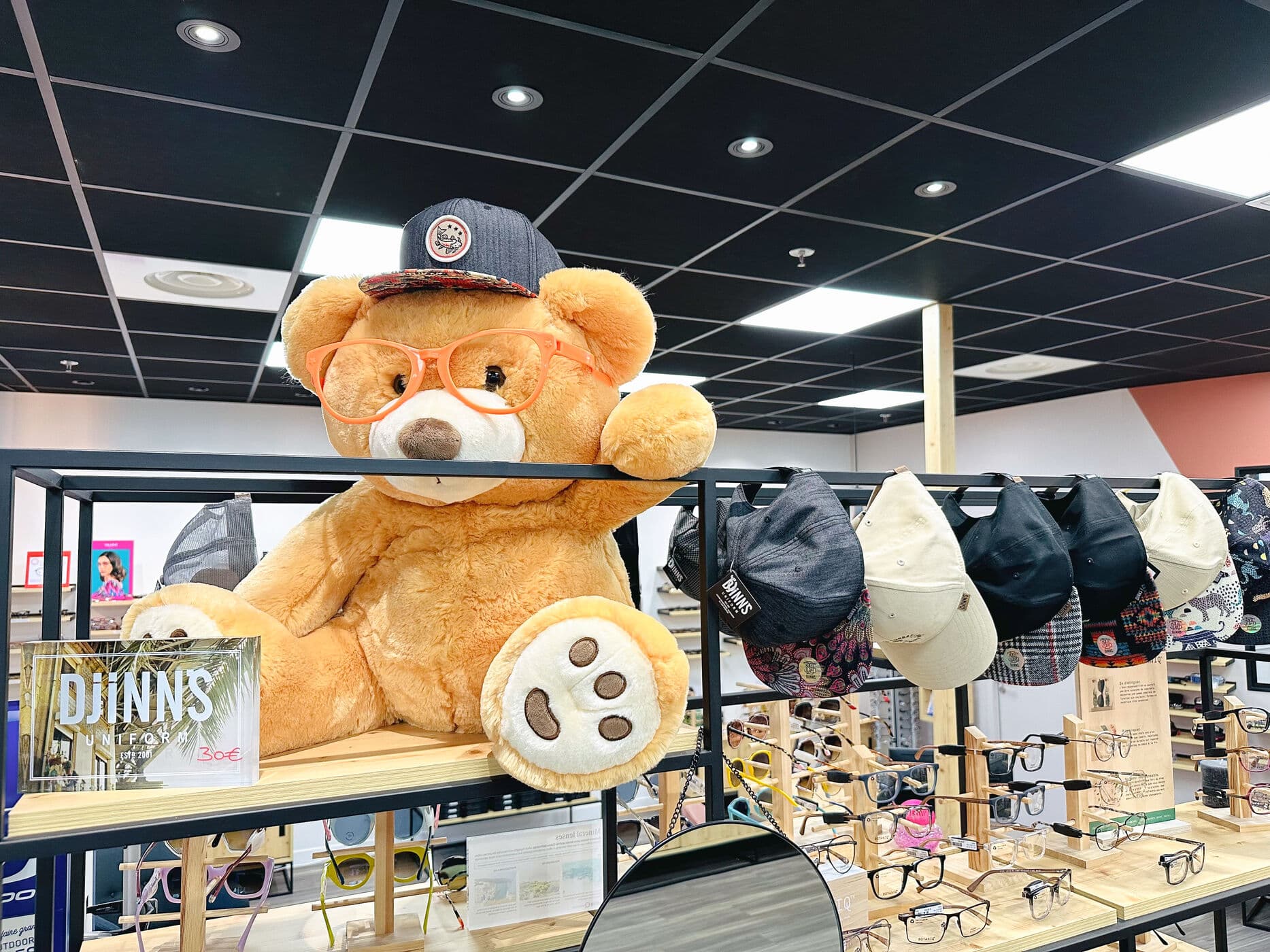 Le grand ours en peluche du magasin, lunettes orange sur le nez, assis parmi les casquettes